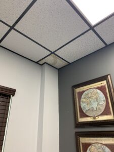 Acoustic drop ceiling tile in a commercial Houston office exhibiting yellow-brown water leak staining near framed wall art, identified during an Aviva Cleaning Services pre-cleaning inspection.